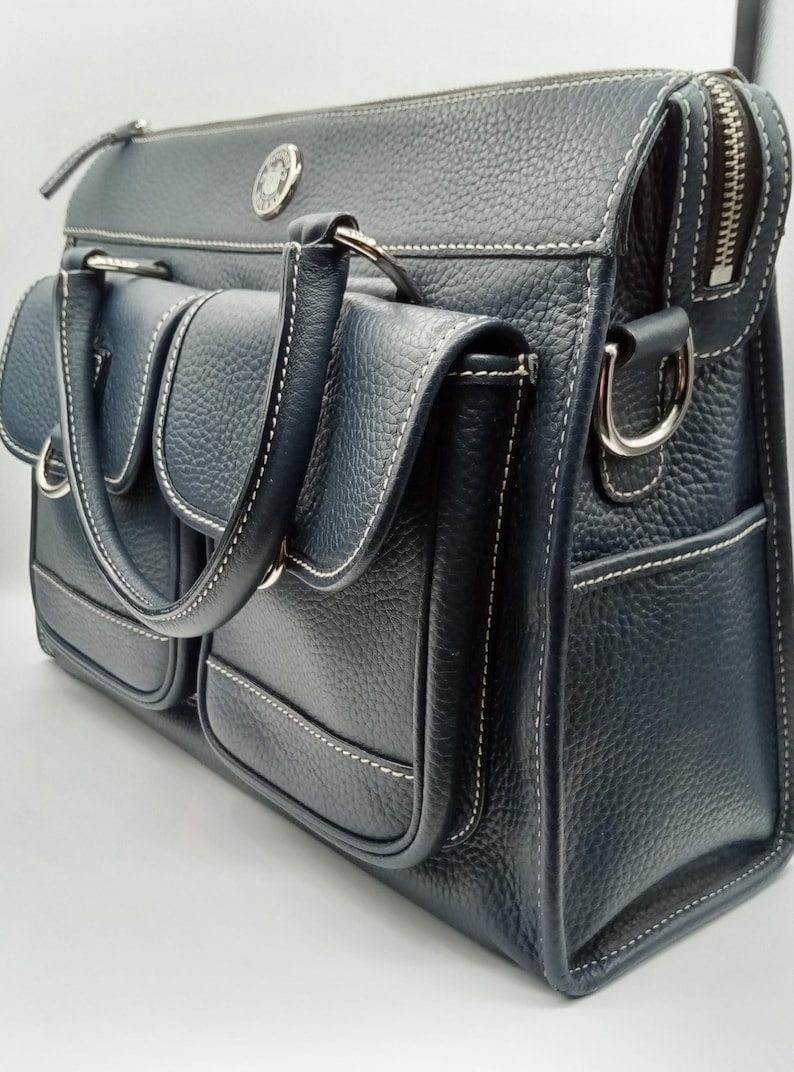 May include: A navy blue leather satchel with white stitching and multiple exterior pockets. The bag features a top zipper closure, silver-tone hardware, and a handle for carrying. A small emblem is visible on the front.