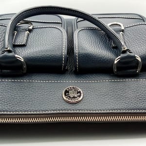 May include: A navy blue leather handbag with two front pockets and a top handle. The bag features white stitching, silver hardware, and a zipper closure. A silver emblem is centered on the front.