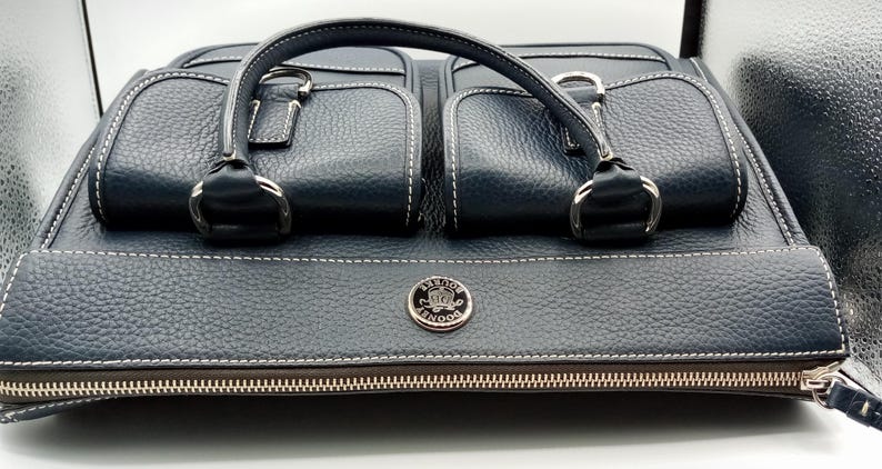 May include: A navy blue leather handbag with two front pockets and a top handle. The bag features white stitching, silver hardware, and a zipper closure. A silver emblem is centered on the front.