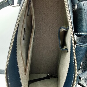 May include: Open view of a navy blue leather handbag with a silver zipper and white stitching. The interior features a beige fabric lining with multiple pockets. The bag has a structured shape and silver hardware.