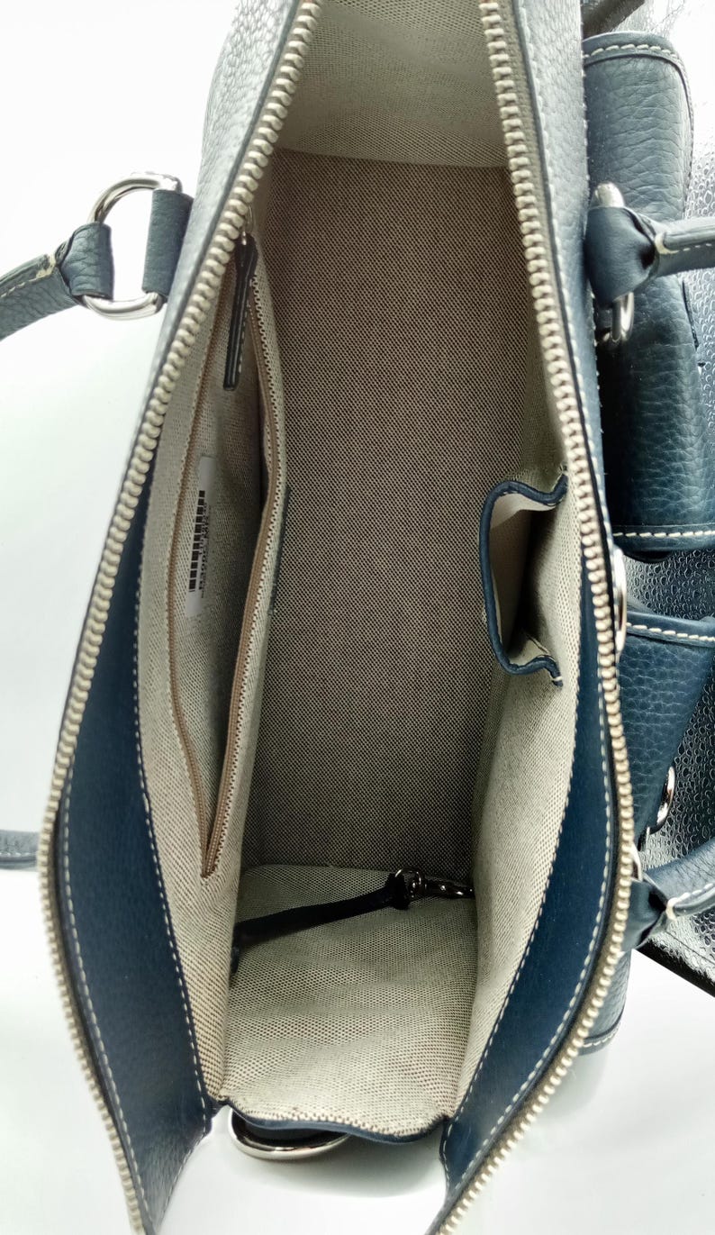 May include: Open view of a navy blue leather handbag with a silver zipper and white stitching. The interior features a beige fabric lining with multiple pockets. The bag has a structured shape and silver hardware.