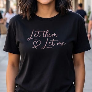 May include: Black t-shirt with the text "Let them ♡ Let me" in a cursive, pink font. The shirt is worn by a person smiling in an outdoor setting. The background includes a sidewalk, buildings, and other people.