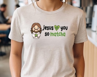 May include: A light beige short-sleeve t-shirt featuring a cartoon illustration of a figure with long brown hair and a beard holding a green cup. The text reads "Jesus [green heart icon] you so matcha" in black and green lettering.