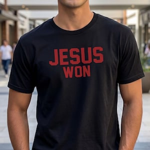 Può includere: T-shirt nero con la scritta rossa "JESUS WON". La maglietta è a girocollo e viene indossata da una persona. Lo sfondo è un centro commerciale con persone e piante.