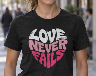 Könnte beinhalten: Ein dunkel anthrazitfarbenes Kurzarm-T-Shirt mit einem herzförmigen Grafikdesign. Der Text lautet LOVE NEVER FAILS mit dem Verweis 1 Corinthians 13:8. Die Schrift verläuft von Weiß im oberen Bereich zu Rosatönen im unteren Bereich.