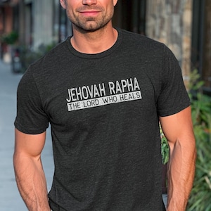 May include: A charcoal gray short-sleeve crew neck t-shirt featuring white distressed-style text on the chest that reads JEHOVAH RAPHA THE LORD WHO HEALS. The shirt has a heathered texture and a standard fit with a simple round neckline.