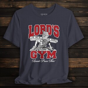Puede incluir: Una camiseta de manga corta gris oscuro con un estampado gr&aacute;fico que incluye el texto rojo "LORD'S GYM" y el texto blanco "THE SIN OF THE WORLD" y "Bench Press This!". La ilustraci&oacute;n muestra una figura haciendo flexiones con una cruz.