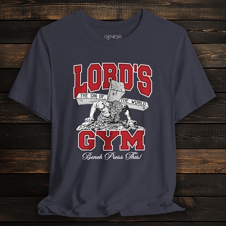 Puede incluir: Una camiseta de manga corta gris oscuro con un estampado gr&aacute;fico que incluye el texto rojo "LORD'S GYM" y el texto blanco "THE SIN OF THE WORLD" y "Bench Press This!". La ilustraci&oacute;n muestra una figura haciendo flexiones con una cruz.