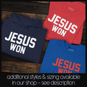 May include: A navy blue hooded sweatshirt, a red t-shirt, and a blue t-shirt, each featuring the white or orange text "JESUS WON" in a bold, block-style font. The garments are displayed together, showcasing the brand name "GENESIS" on the inner labels.