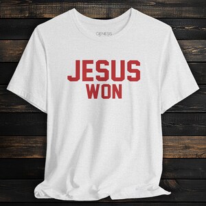 May include: A white short-sleeved t-shirt featuring the text JESUS WON in bold red block letters across the chest. The shirt has a crew neckline and a small Genesis brand logo printed in grey on the inside of the collar area.
