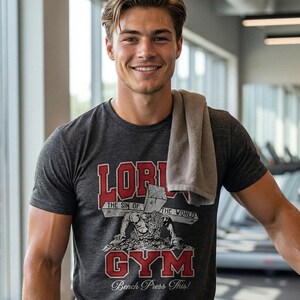 Puede incluir: Una camiseta de manga corta gris carb&oacute;n con un estampado gr&aacute;fico que incluye el texto rojo "LORD" y "GYM", el texto blanco "THE SIN OF THE WORLD" y "Bench Press This!" y una ilustraci&oacute;n de una figura cargando una cruz de madera.