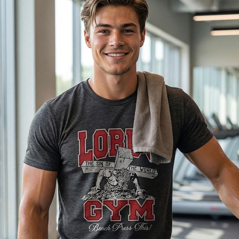 Puede incluir: Una camiseta de manga corta gris carb&oacute;n con un estampado gr&aacute;fico que incluye el texto rojo "LORD" y "GYM", el texto blanco "THE SIN OF THE WORLD" y "Bench Press This!" y una ilustraci&oacute;n de una figura cargando una cruz de madera.