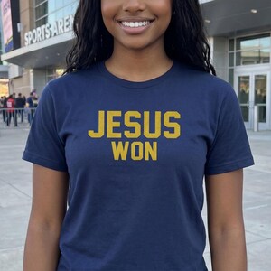 May include: A navy blue short-sleeved t-shirt featuring the text "JESUS WON" printed in a bold, gold-colored collegiate-style font across the chest. The shirt has a classic crew neckline and a standard fit, providing a simple and direct design.