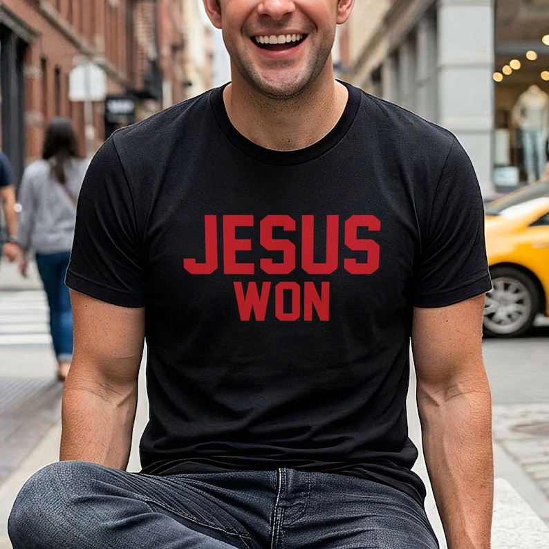 May include: A black short-sleeved t-shirt featuring the text JESUS WON printed in bold red capital letters across the chest. The garment has a classic crew neckline and a standard fit, providing a simple and direct design for casual wear.
