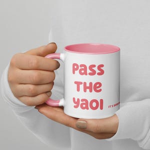 Puede incluir: Taza de cerámica blanca con interior y asa rosas. La taza presenta el texto "Pass the Yaoi" en rosa, con un texto más pequeño "It's been" en el lateral. La taza está sujeta por una persona.