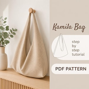 以下が含まれることがあります： 白い壁の木製ペグにベージュ色のKamilaバッグが吊るされています。バッグは、長いストラップが付いた、大きく開いたデザインです。右側のテキストは「Kamila Bag」、「step by step tutorial」、「PDF PATTERN」と書かれています。