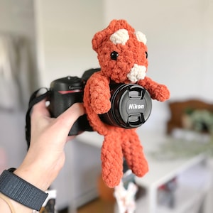 May include: A crocheted orange and white plush toy with black eyes is attached to a black camera lens with the word "Nikon" printed on it.