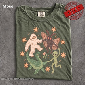 May include: A moss green t-shirt featuring a graphic print of a white yeti, a brown moth, a green dinosaur, and a green alien holding hands in a circle. The design includes orange stars and a small flower, with a Comfort Colors brand label at the neck.