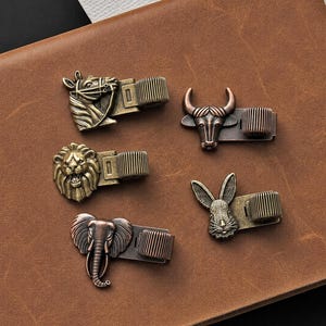 May include: A set of five metal pen holders featuring animal heads including a horse, a bull, a lion, a rabbit, and an elephant. Each holder has a metallic finish in bronze or copper tones and includes a cylindrical loop for securing a writing instrument.