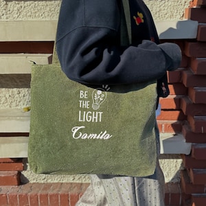 May include: Olive green corduroy tote bag with the text "BE THE LIGHT" and a lightbulb graphic. The bag has a long shoulder strap and a zipper closure. The bag is being carried in front of a brick wall.