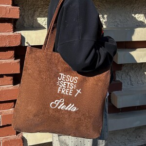 May include: A brown corduroy tote bag with the text "JESUS SETS FREE" and a cross graphic in white. The name "Stella" is embroidered below. The bag has a zipper closure and long straps.