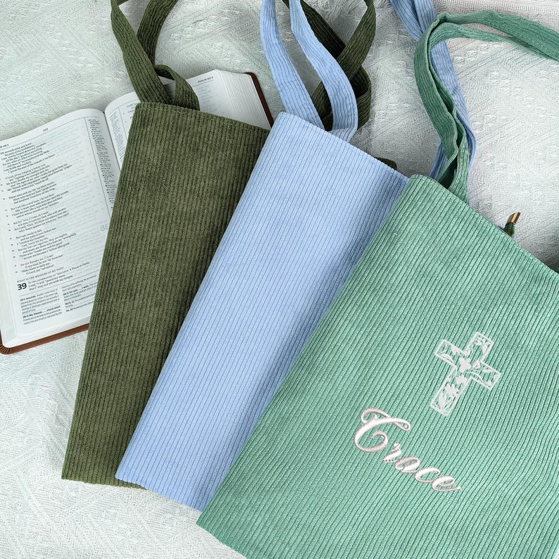 May include: Three corduroy tote bags in olive green, light blue, and seafoam green. The seafoam green bag has a white embroidered cross and the word "Grace". Each bag has matching corduroy straps and is next to an open book.