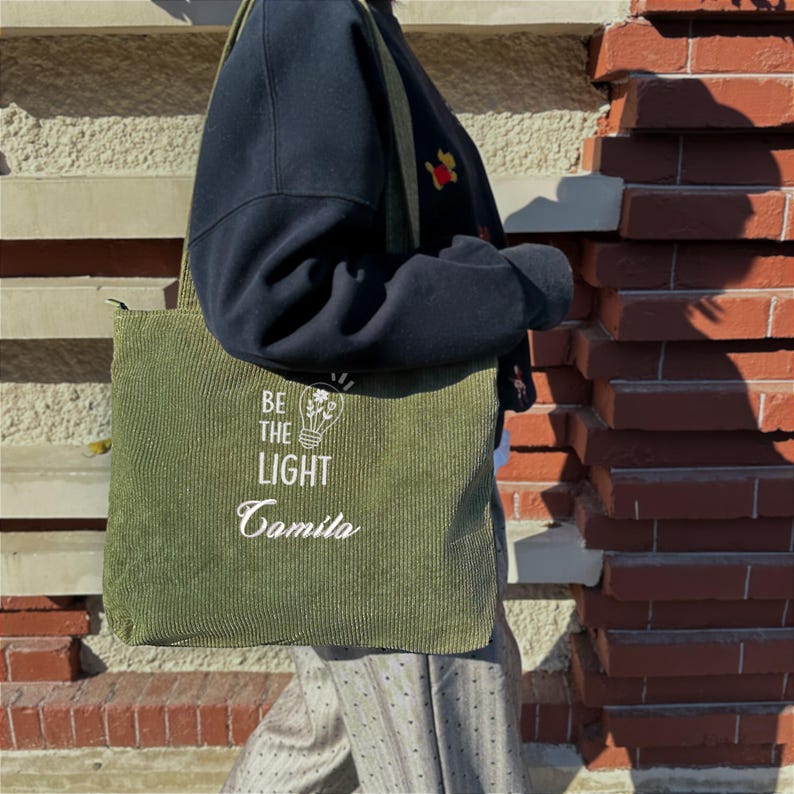 May include: Olive green corduroy tote bag with the text "BE THE LIGHT" and a lightbulb graphic. The bag has a long shoulder strap and a zipper closure. The bag is being carried in front of a brick wall.