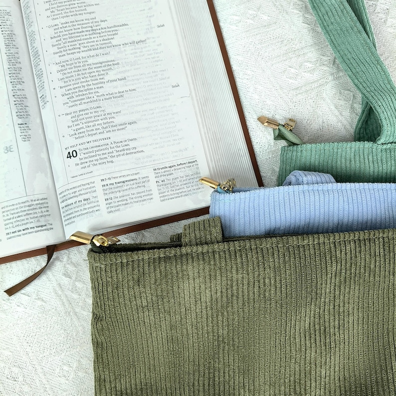 May include: An open Bible rests beside a green corduroy tote bag with a gold zipper. A light blue corduroy pouch and a green corduroy strap are also visible. The pages of the Bible are open to a passage with the number 40.