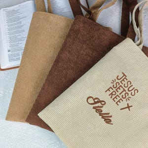 May include: Three corduroy tote bags in tan, brown, and cream colors. The cream bag has the embroidered text "JESUS SETS FREE" and the name "Stella". The bags have matching corduroy handles.