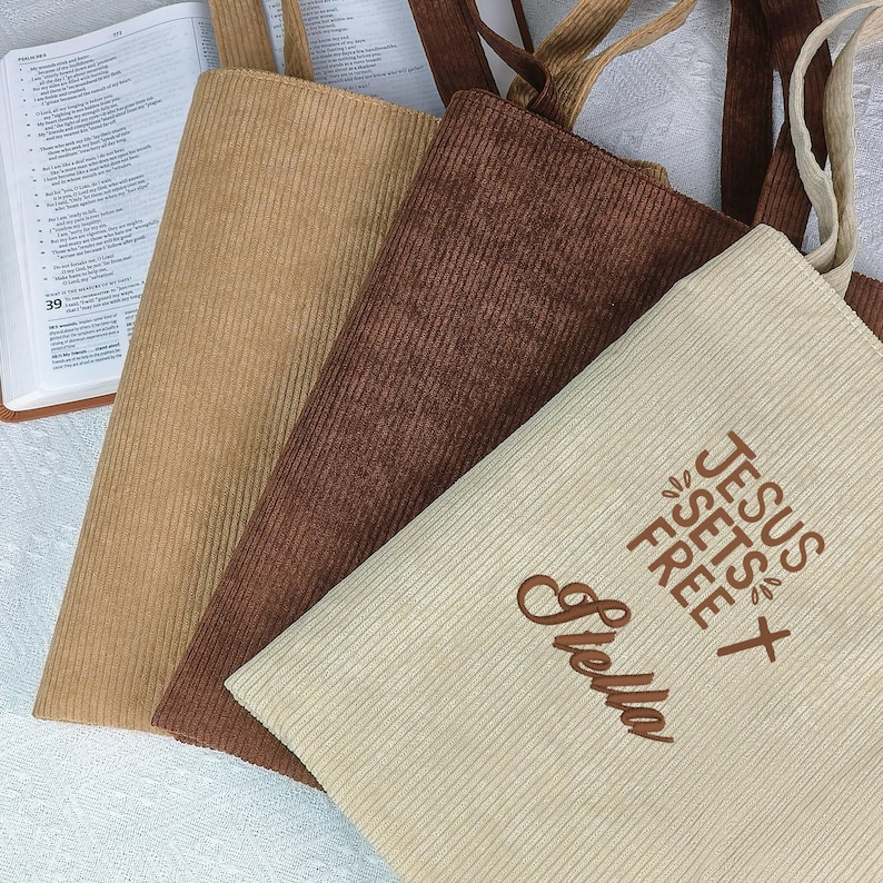 May include: Three corduroy tote bags in tan, brown, and cream colors. The cream bag has the embroidered text "JESUS SETS FREE" and the name "Stella". The bags have matching corduroy handles.
