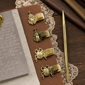 May include: Four gold-toned metal pen clips, each featuring a distinct beetle design and an attached spring-style pen holder. The clips are arranged vertically on a brown leather surface next to a slim gold-toned metal ballpoint pen.
