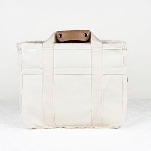 Op de afbeelding: Cr&egrave;mekleurige canvas draagtas met stevige handvatten en een bruine leren handgreep. De tas heeft meerdere vakken en versterkte stiksels. Een praktische en stijlvolle accessoire voor dagelijks gebruik.