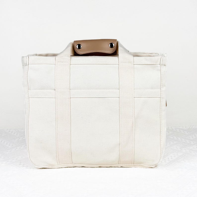 Op de afbeelding: Cr&egrave;mekleurige canvas draagtas met stevige handvatten en een bruine leren handgreep. De tas heeft meerdere vakken en versterkte stiksels. Een praktische en stijlvolle accessoire voor dagelijks gebruik.