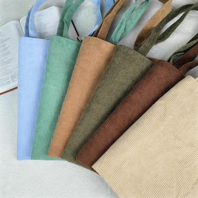 May include: A collection of corduroy tote bags in various colors, including light blue, green, tan, olive green, brown, and cream. Each bag has a matching corduroy handle. The bags are arranged in a diagonal pattern.