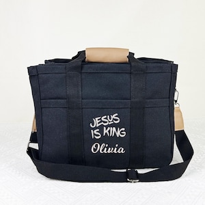 Op de afbeelding: Zwarte tote bag met meerdere vakken en een schouderriem. De tas heeft de geborduurde tekst "JESUS IS KING Olivia" in het wit. Het heeft bruin getinte handvatten en accenten.