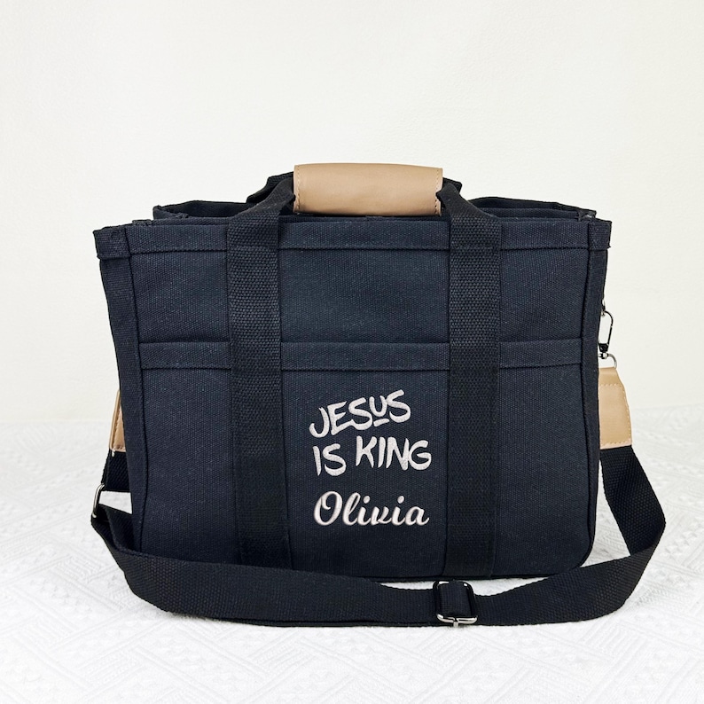 Op de afbeelding: Zwarte tote bag met meerdere vakken en een schouderriem. De tas heeft de geborduurde tekst "JESUS IS KING Olivia" in het wit. Het heeft bruin getinte handvatten en accenten.