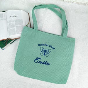 May include: A light green corduroy tote bag with matching handles. The bag features the text "Rooted in Christ" above a butterfly graphic and the name "Emilia" in navy blue. An open book with the word "Samuel" is visible.