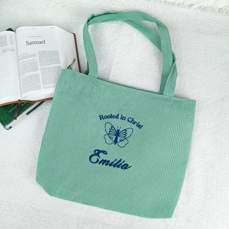May include: A light green corduroy tote bag with matching handles. The bag features the text "Rooted in Christ" above a butterfly graphic and the name "Emilia" in navy blue. An open book with the word "Samuel" is visible.