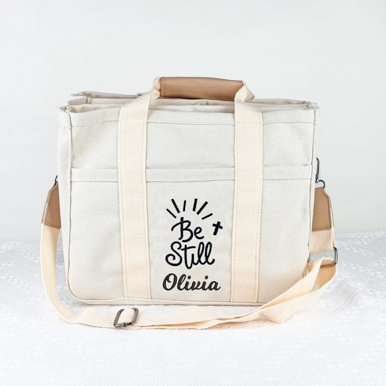 May include: Cream-colored tote bag with a shoulder strap and handles. The bag features the text "Be Still Olivia" in black script. The bag has a rectangular shape with multiple pockets and a light brown leather handle.