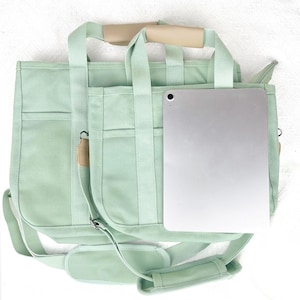Op de afbeelding: Een lichtgroene canvas draagtas met meerdere vakken en een afneembare schouderriem. De tas heeft een beige handvat en een zilveren tablet is erin geplaatst. De tas is ontworpen voor het dragen van items.