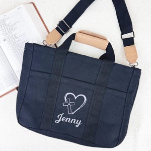 Op de afbeelding: Een marineblauwe draagtas met een wit geborduurd hart- en kruisontwerp en de naam "Jenny". De tas heeft zwarte banden, een bruin handvat en een afneembare schouderriem. Op de achtergrond is een open boek te zien.
