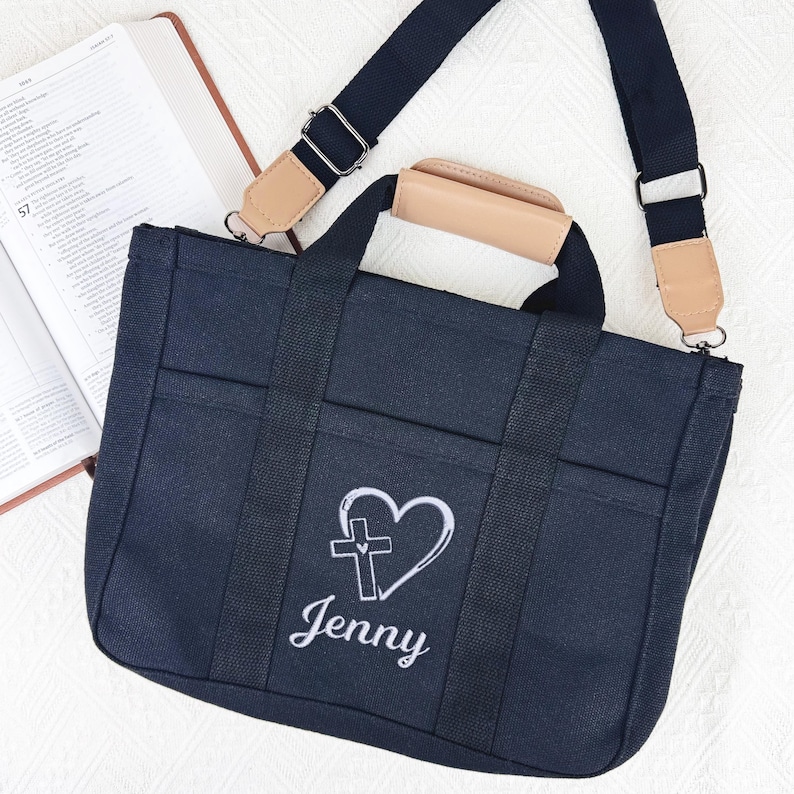 Op de afbeelding: Een marineblauwe draagtas met een wit geborduurd hart- en kruisontwerp en de naam "Jenny". De tas heeft zwarte banden, een bruin handvat en een afneembare schouderriem. Op de achtergrond is een open boek te zien.