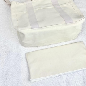 Op de afbeelding: Off-white canvas draagtas met bijpassend etui. De tas heeft een rechthoekige vorm met een versterkte bodem en handvatten aan de bovenkant. Het etui is rechthoekig en lijkt van hetzelfde materiaal te zijn gemaakt. De tas ligt op een wit, getextureerd oppervlak.