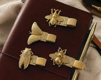 May include: Four gold-toned metal pen holders featuring insect and butterfly motifs. Each holder has a decorative top piece shaped like a bee, a butterfly, or a cicada, attached to a flexible, ribbed metal loop designed to secure a writing instrument.