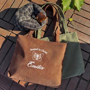 May include: A brown corduroy tote bag with the text "Rooted in Christ" and the name "Emilia" in white. A green corduroy tote bag is next to it. A plaid hat with a button and a leaf is on top of the bags.