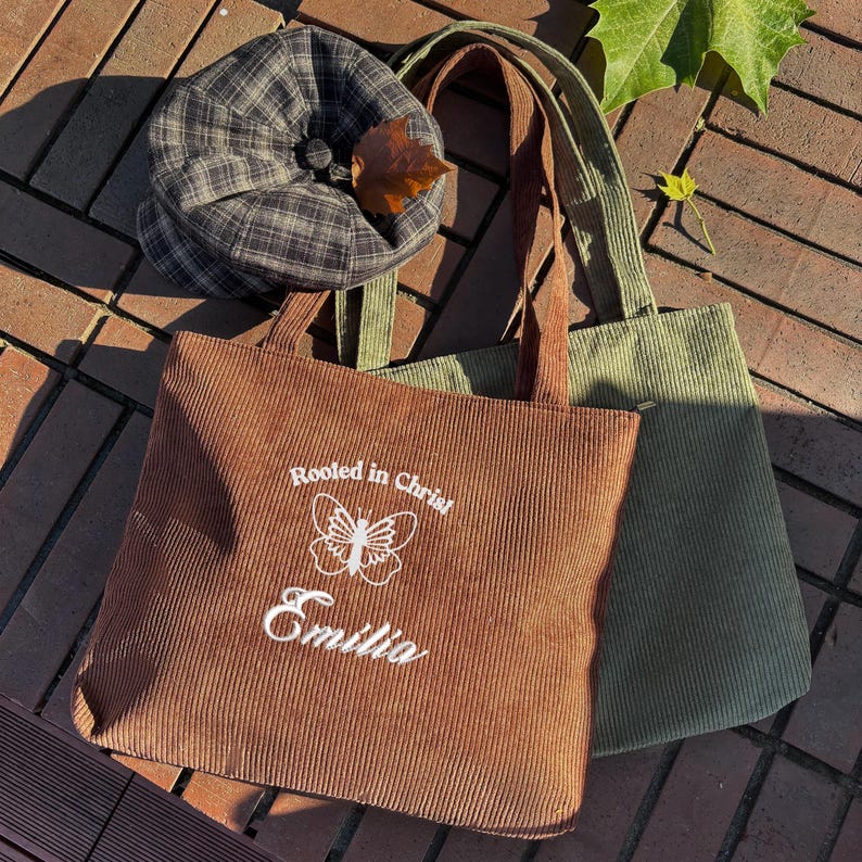 May include: A brown corduroy tote bag with the text "Rooted in Christ" and the name "Emilia" in white. A green corduroy tote bag is next to it. A plaid hat with a button and a leaf is on top of the bags.