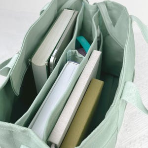 Op de afbeelding: Een lichtgroene canvas draagtas met meerdere binnenvakken. De tas is gevuld met boeken, een tablet en andere kleine items. De tas heeft twee handvatten en is gemaakt van een duurzame stof.