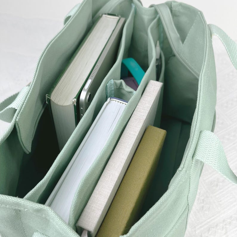 Op de afbeelding: Een lichtgroene canvas draagtas met meerdere binnenvakken. De tas is gevuld met boeken, een tablet en andere kleine items. De tas heeft twee handvatten en is gemaakt van een duurzame stof.