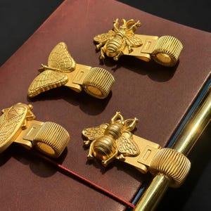 May include: Four gold-toned metal pen holders featuring insect and butterfly designs. Each holder has a textured, cylindrical loop for a pen and a flat clip base. The designs include two bees, one butterfly, and one cicada, all with a matte finish.