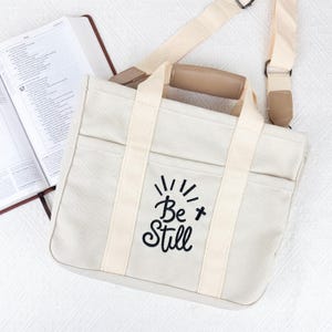 Op de afbeelding: Cr&egrave;me canvas draagtas met de geborduurde tekst "Be Still" in zwart. De tas heeft twee handvatten en een afneembare schouderriem. Op de achtergrond is een open boek zichtbaar.